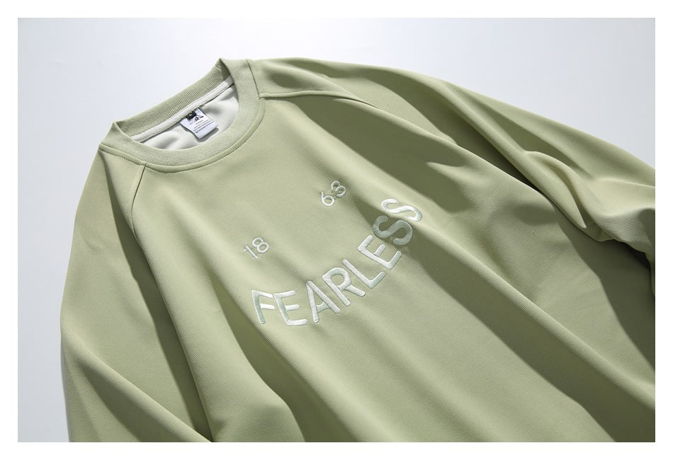 Close-up of a sage-green sweatshirt chest with white embroidered “FEARLESS” lettering and small embroidered symbols.