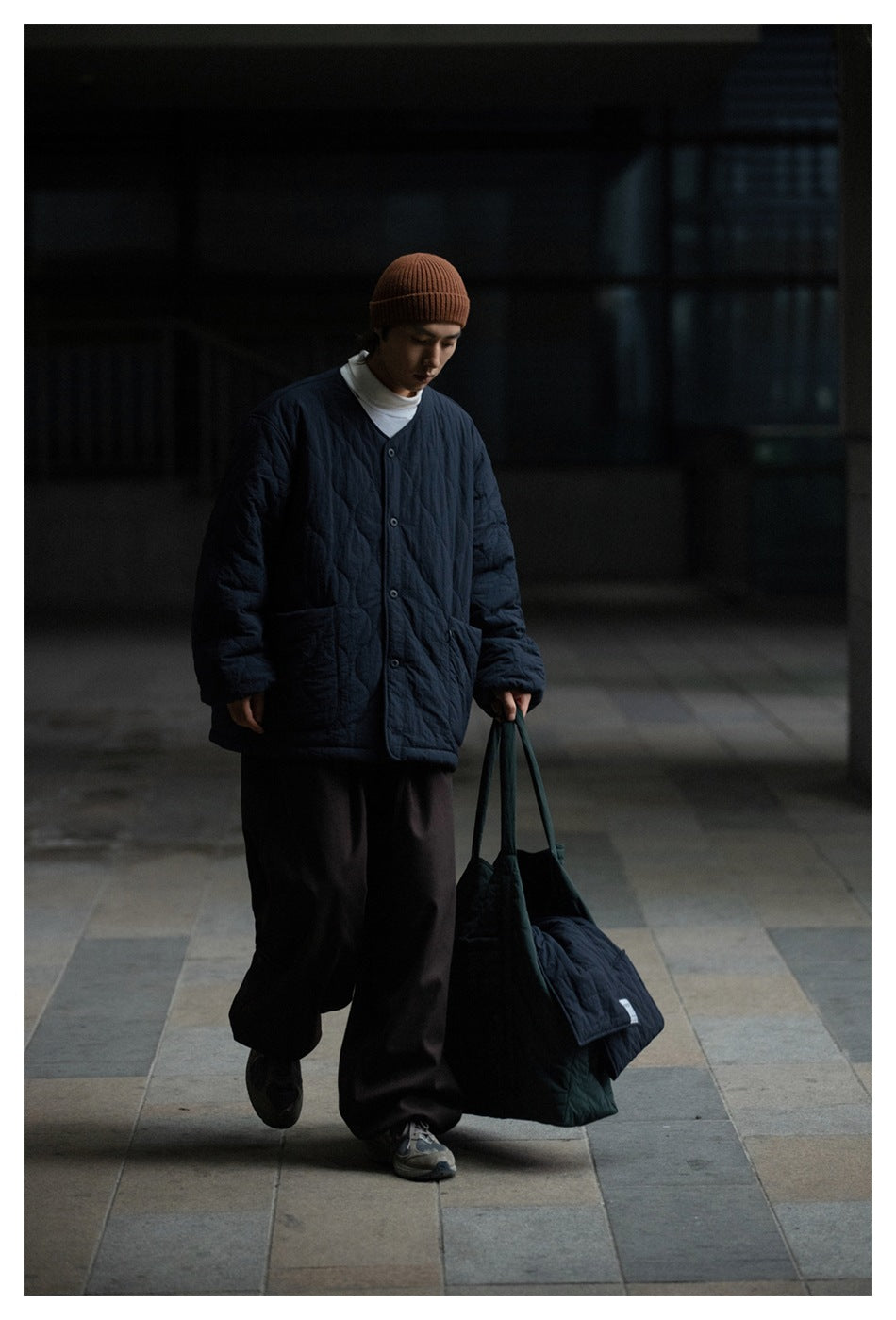Model walking in a dark navy quilted button-front jacket with patch pockets and a rust beanie.