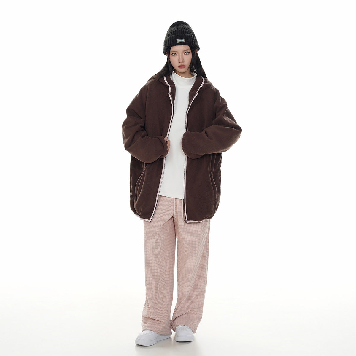 Model wearing an oversized dark brown hooded zip jacket with white trim over a white top and pale pink checked trousers.