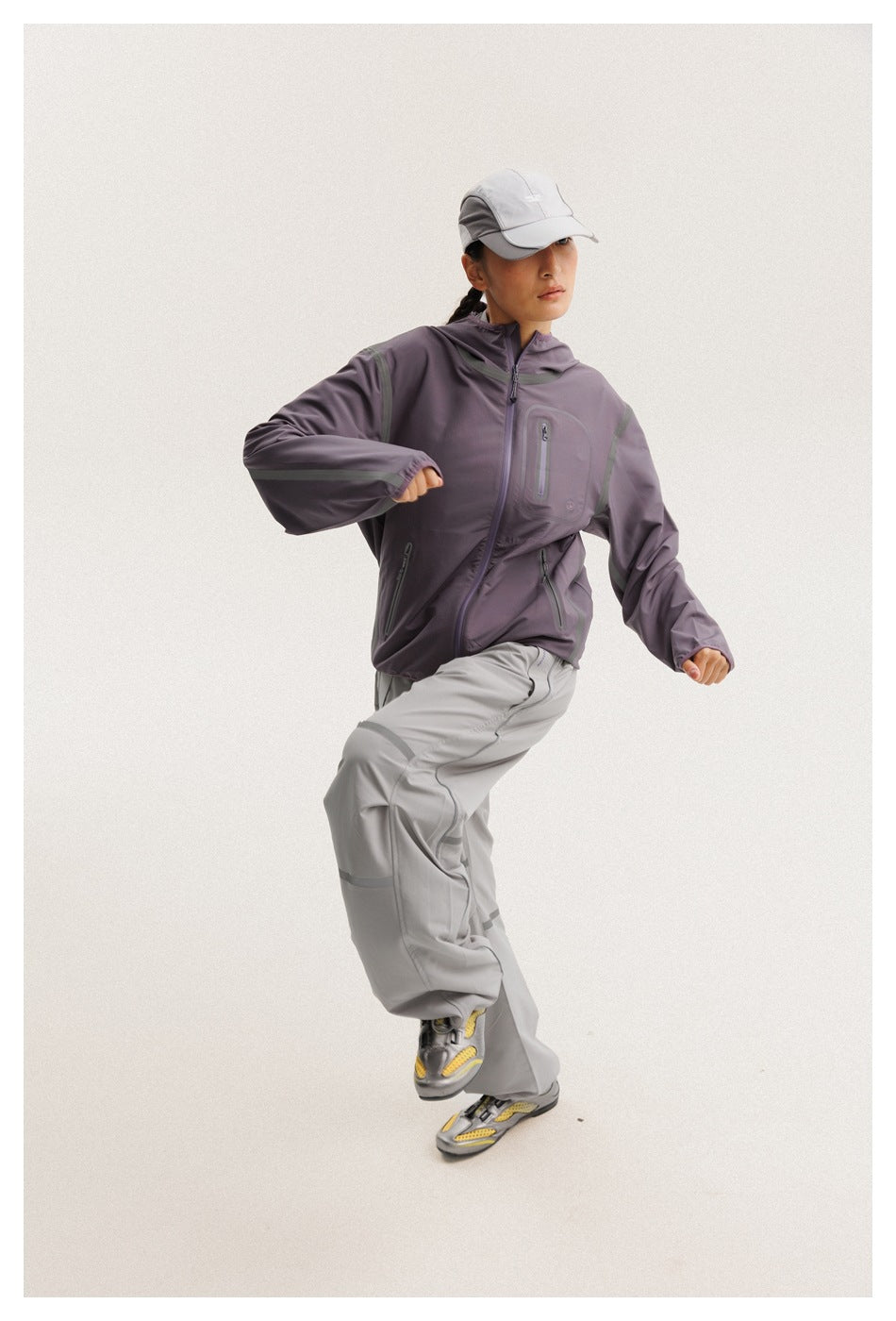 Model wearing a muted purple zip-up hooded jacket, light gray pants, and a gray cap.