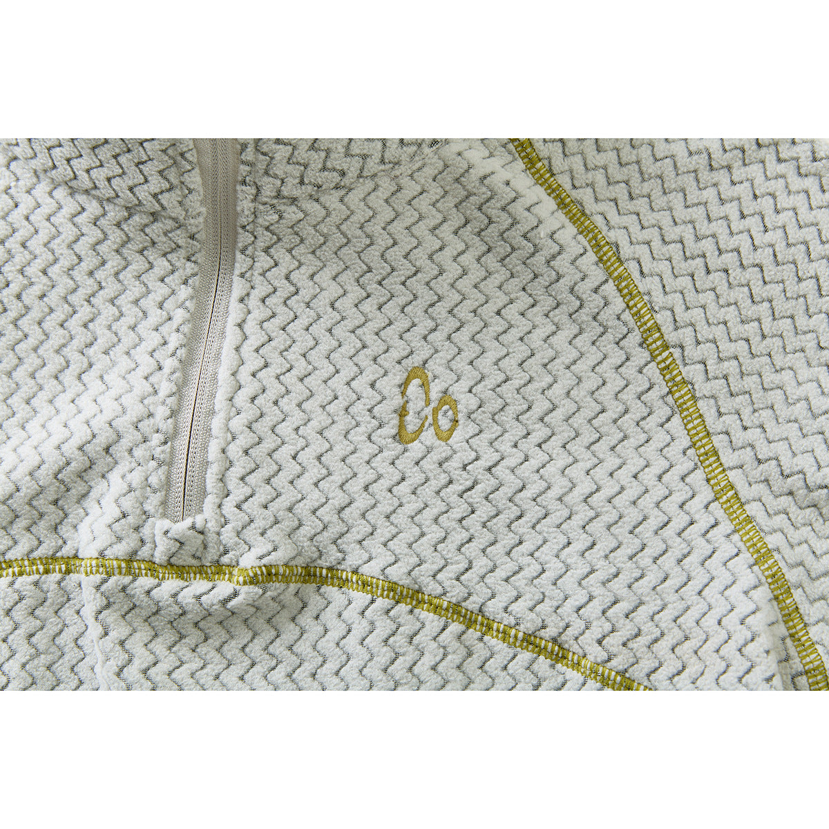 Close-up of a cream and gray zigzag-patterned top with a small gold embroidered chest emblem and mustard stitching.