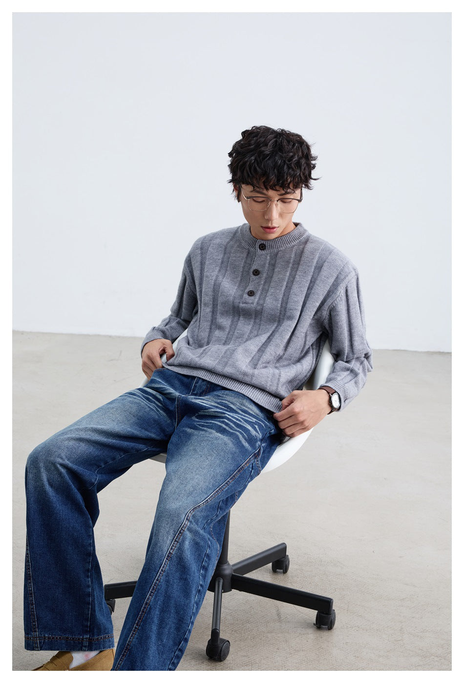 Model seated in a gray sweater with vertical panels and a three-button neck, paired with blue jeans.
