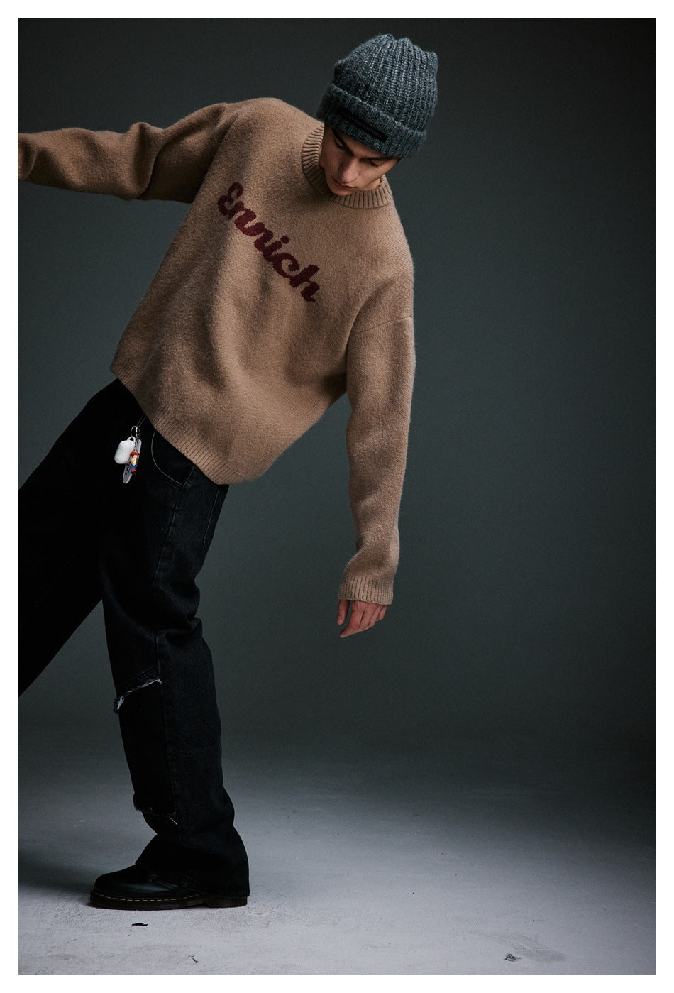 Model leaning in a taupe sweater with burgundy chest lettering, black jeans, and a charcoal beanie.