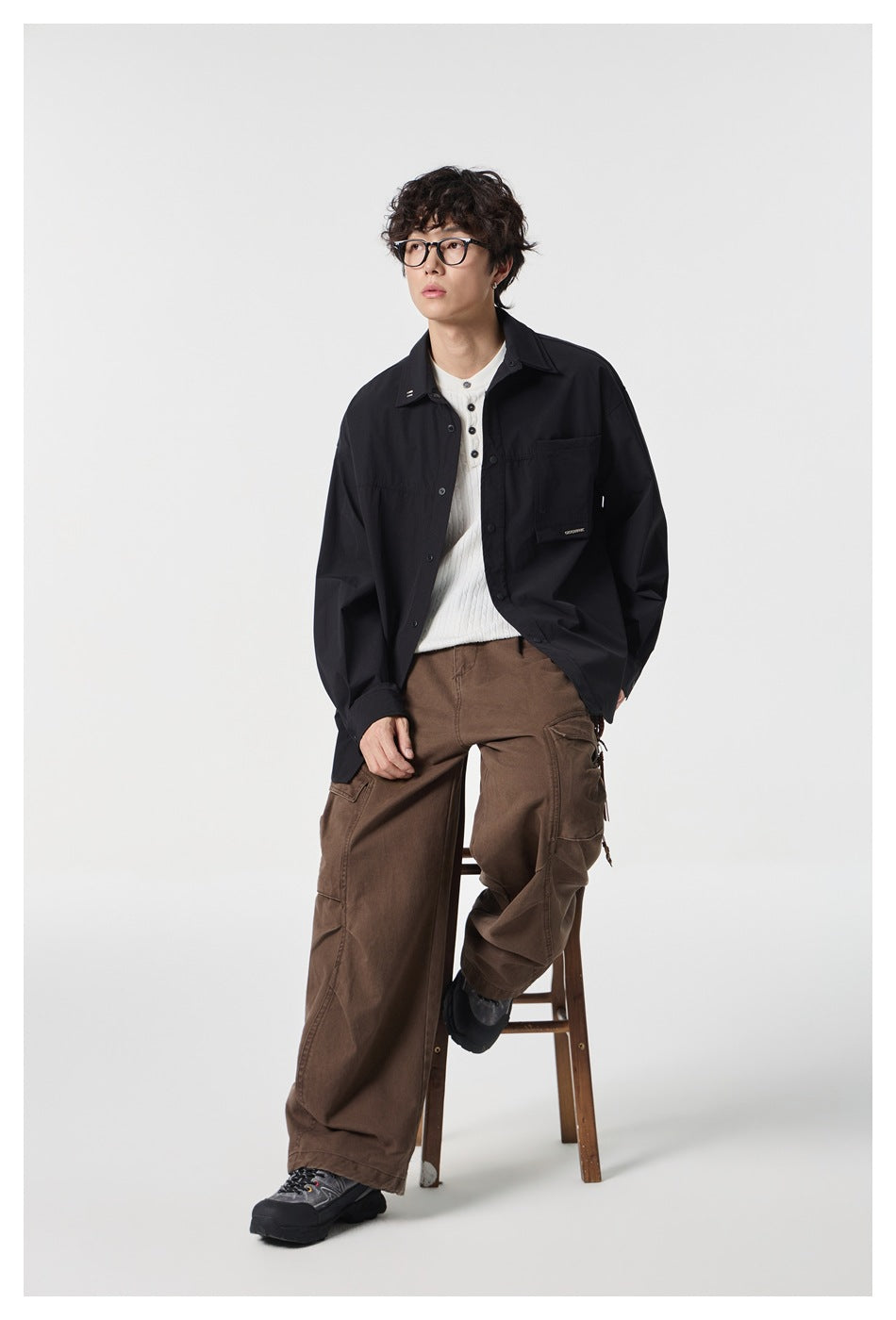 Model seated in an open black button-front shirt over a white knit top and brown cargo trousers.