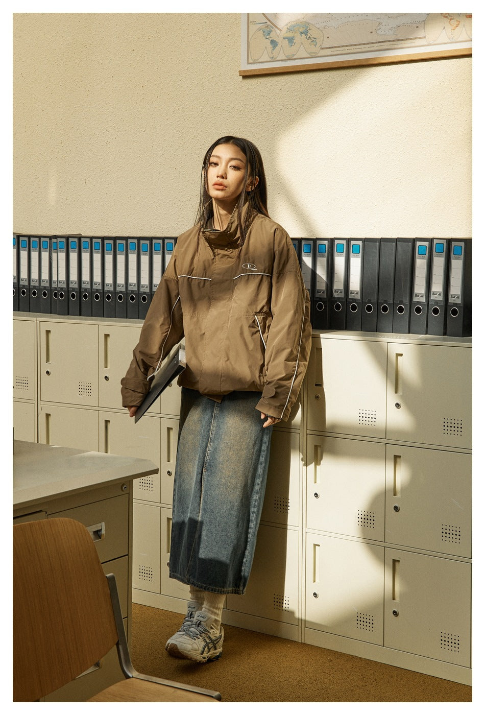 Person leaning on filing cabinets wearing a brown piped jacket with a long denim skirt and sneakers