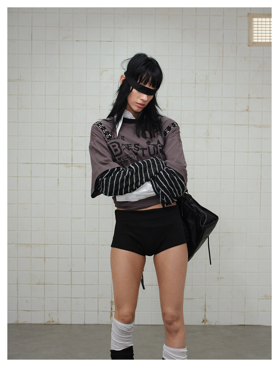 Model wearing a gray graphic top with striped sleeves and black shorts, standing with arms crossed.