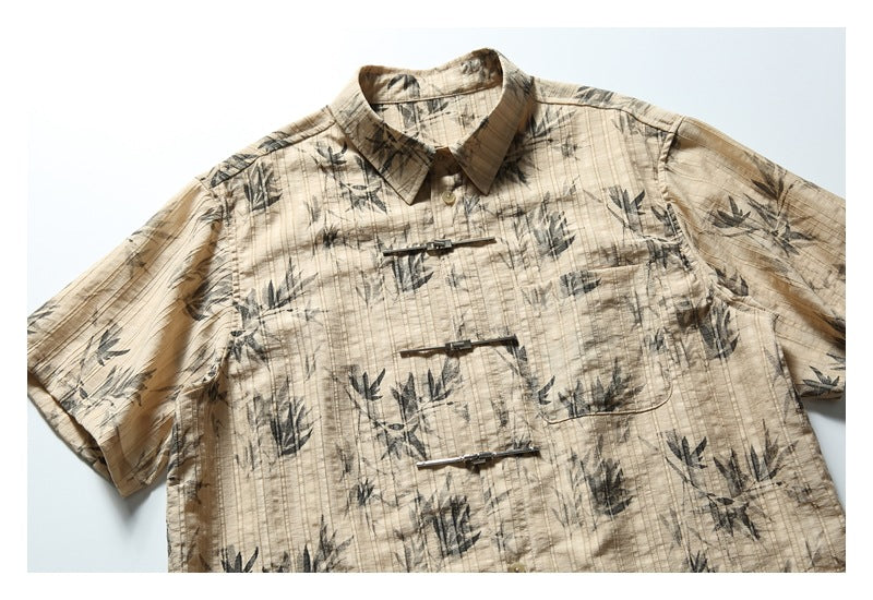 Front view of a tan short-sleeve collared shirt with dark botanical print, a chest pocket, and three silver clasps.