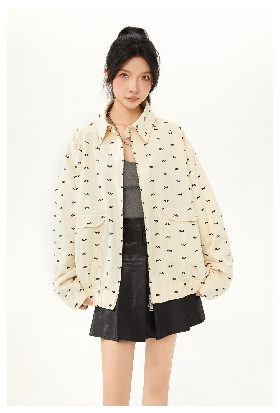 Front view of a model wearing a cream, black-bow-print jacket with flap pockets over a gray top and black skirt.