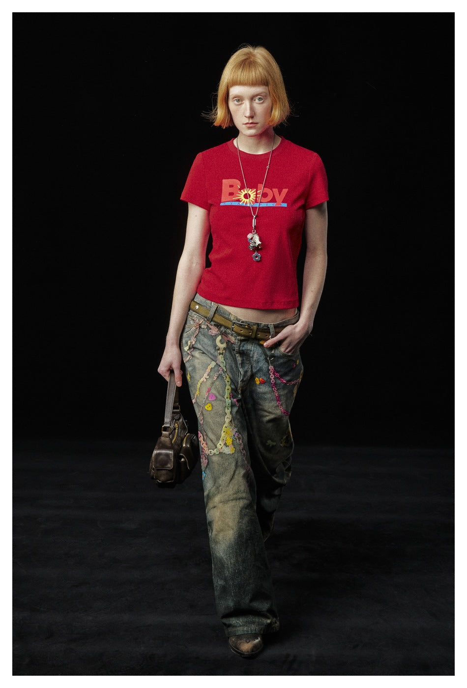 Full-length model wearing a red graphic T-shirt and embellished loose jeans against a black background.