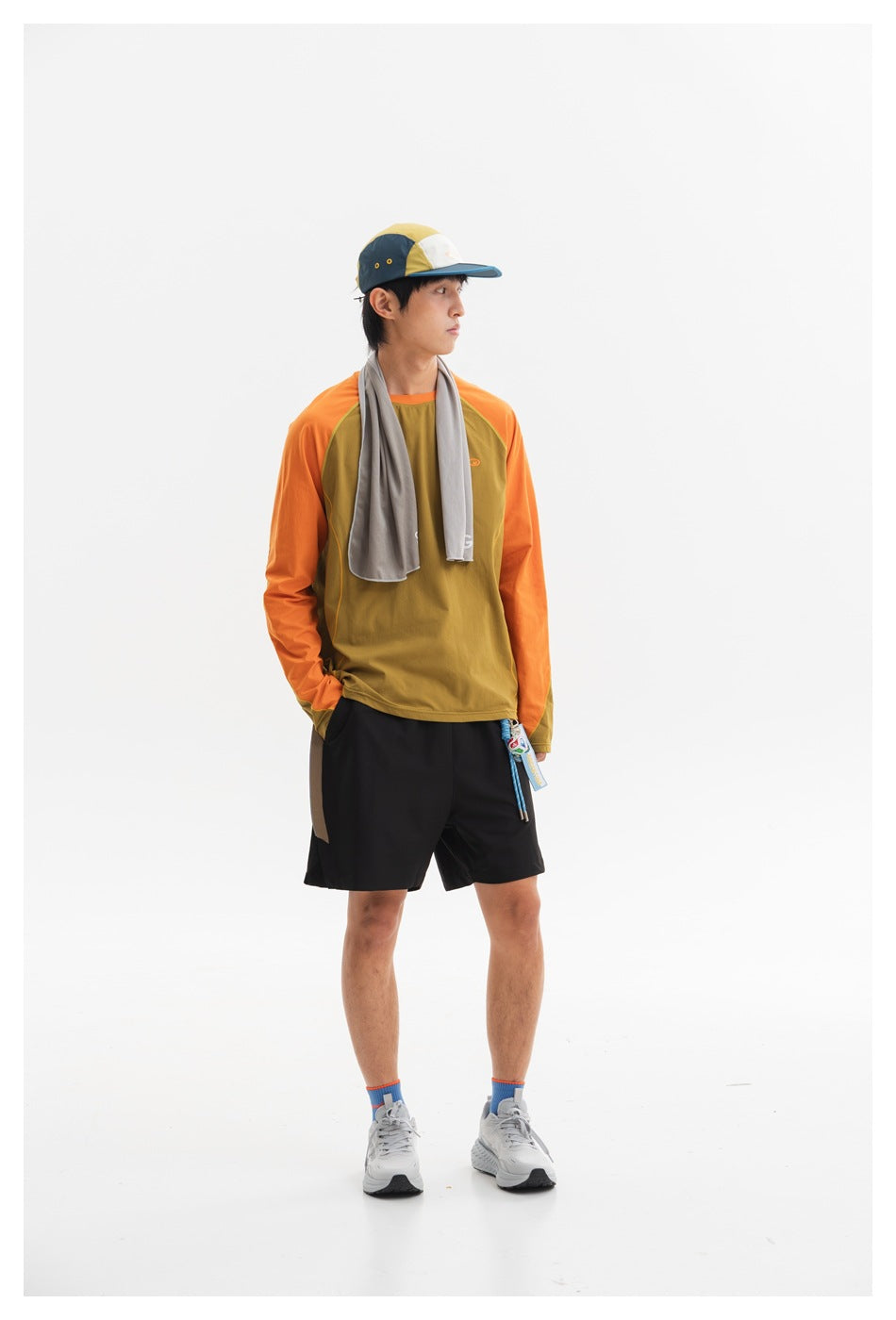 Full-length front view of a model in an olive top with orange sleeves, black shorts, and a color-block cap.