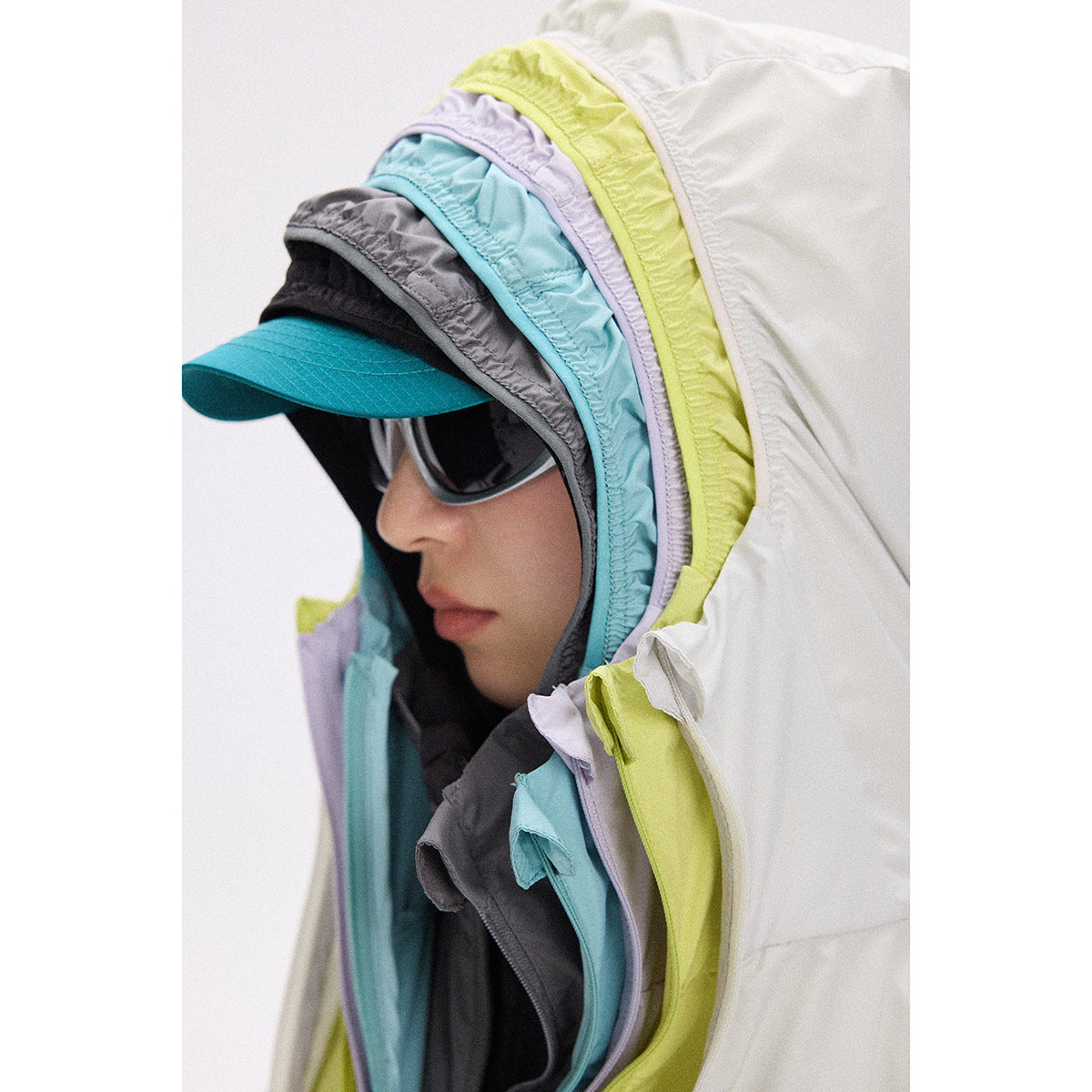 Close-up of a person wearing sunglasses and a cap beneath layered white, lime, lavender, aqua, and gray hoods.