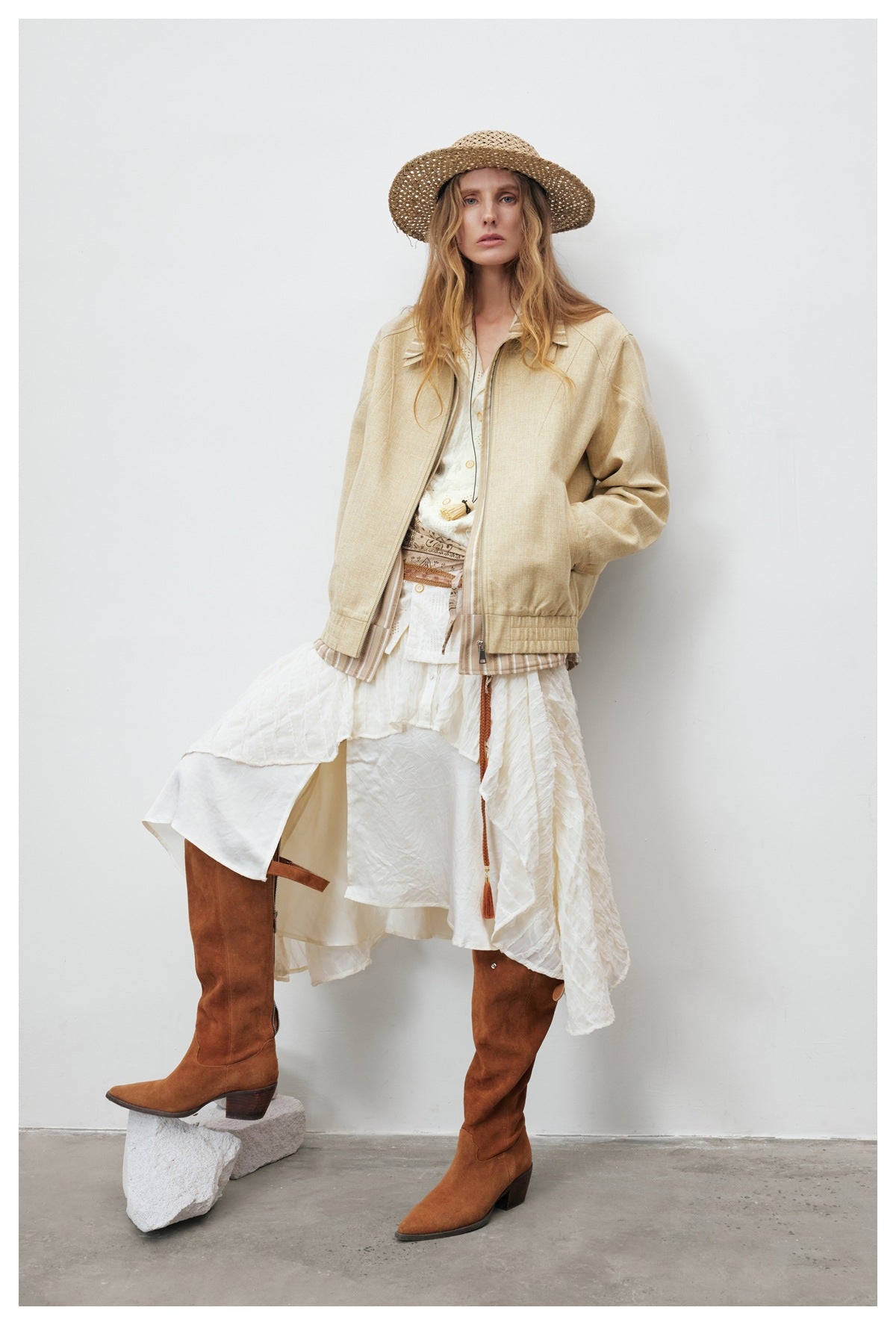 Model wearing a pale beige zip jacket over a layered white skirt, with tall brown boots and a woven hat.