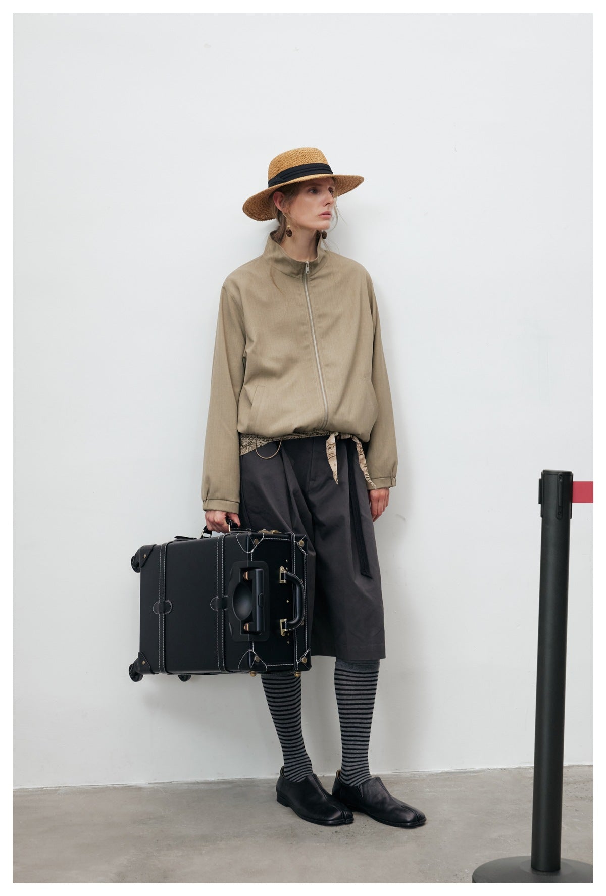 Model wearing a taupe zip-front jacket with a stand collar, styled with a dark skirt, striped socks and a straw hat.