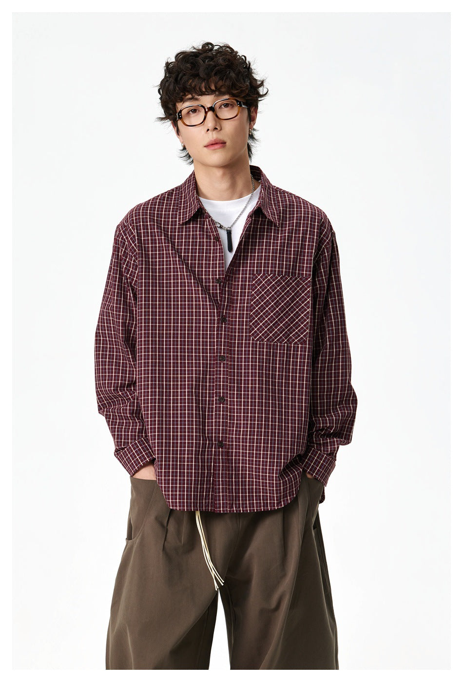 Front view of a model wearing a burgundy plaid button-up shirt with a contrasting chest pocket and loose brown trousers.