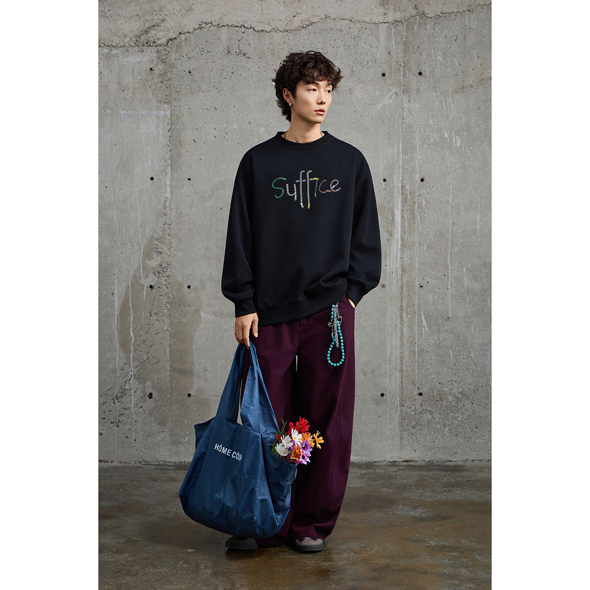 Full-length model in a black crewneck with multicolored lettering and loose plum trousers, holding a blue tote with flowers.