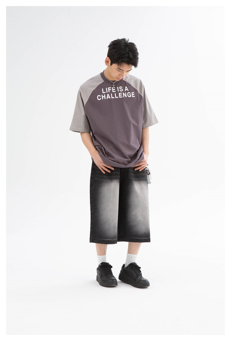 Model looking down, wearing a purple-gray oversized raglan T-shirt with slogan print, black faded shorts and sneakers
