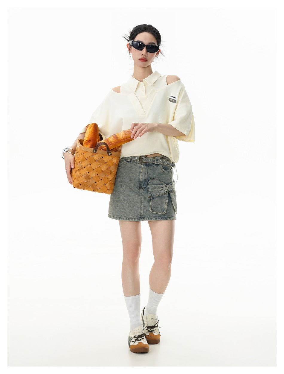 Model wearing a cream cropped polo top with shoulder cutouts, denim bow skirt, holding a woven basket with bread