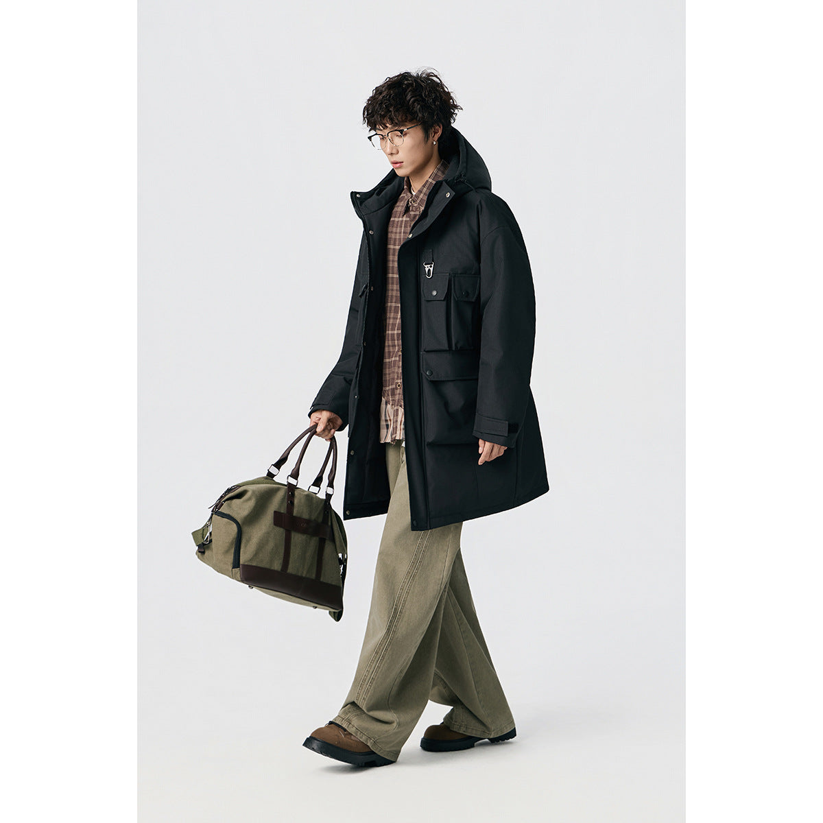 Model walking in a black hooded long coat with flap pockets, plaid shirt and loose khaki trousers, carrying a duffel bag.