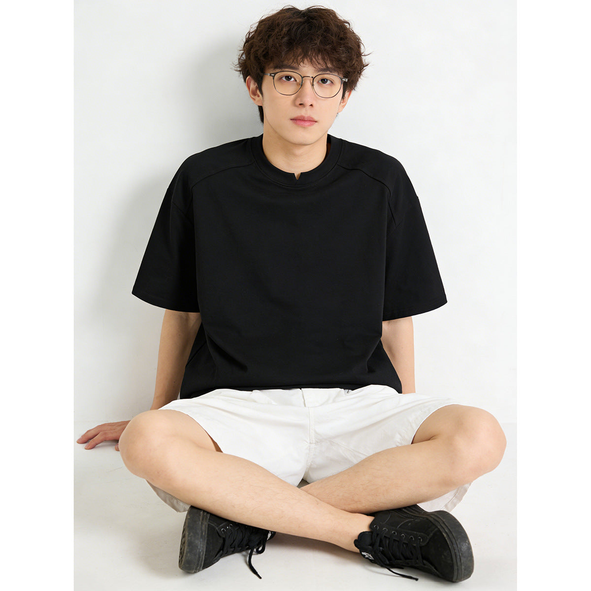 Seated model wearing a plain black short-sleeve T-shirt with a small center neckline notch and white shorts.