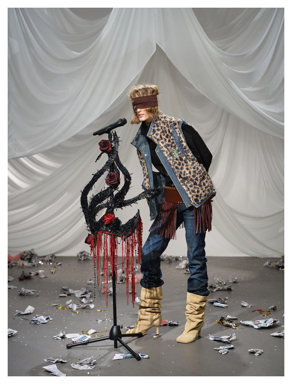 Model leans beside a microphone in a leopard-print vest with denim trim and long burgundy fringe, jeans and tan boots.