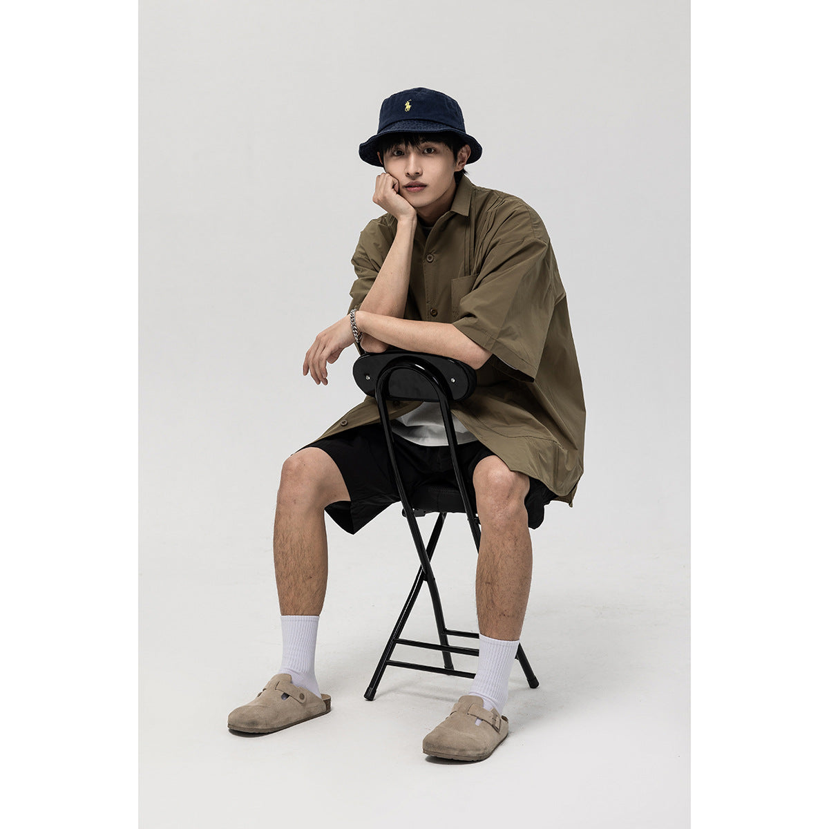 Model seated in an olive short-sleeve button-up shirt with black shorts and a navy bucket hat, shown from the front.