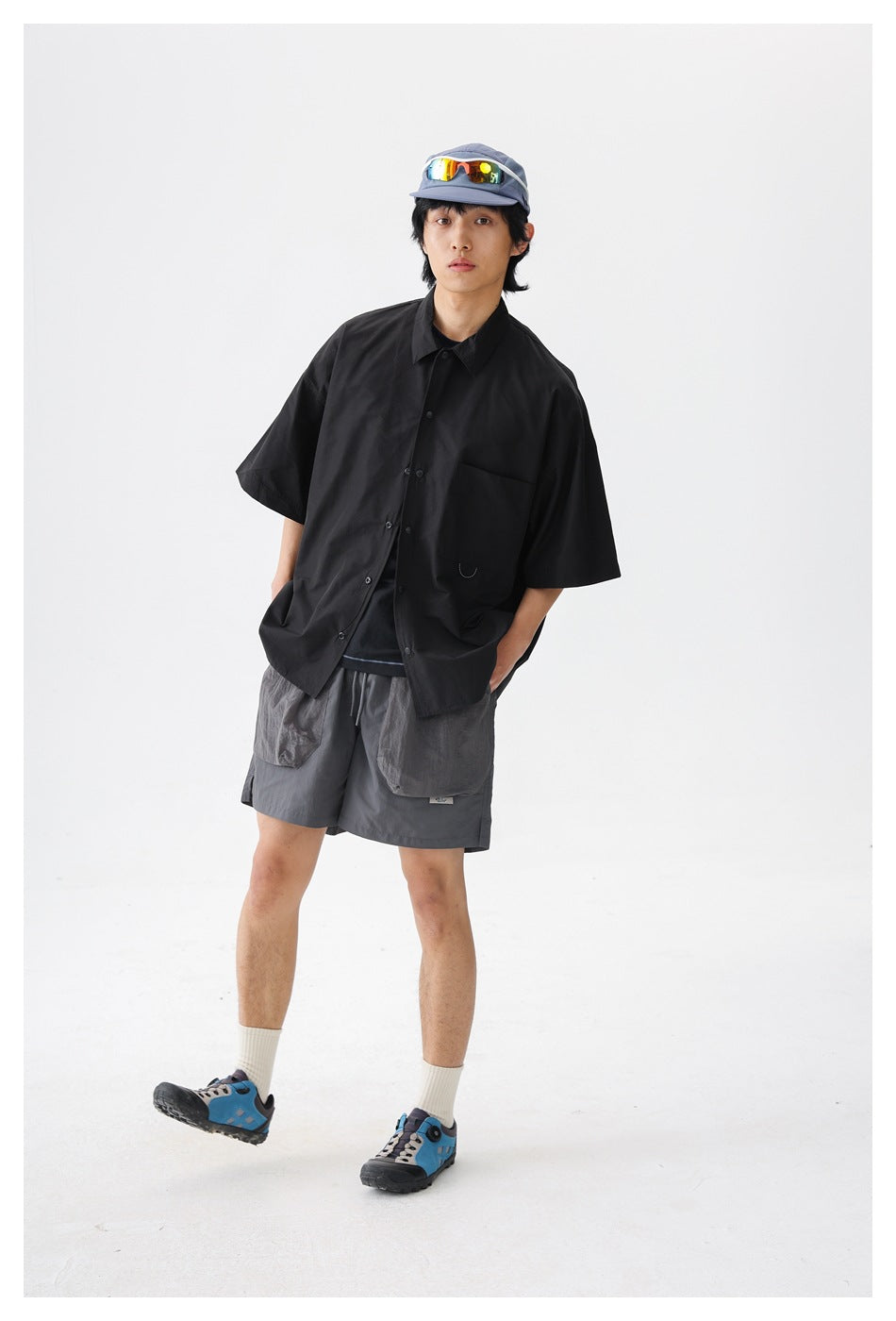 Model wearing an oversized black short-sleeve button-up shirt and gray shorts in a white studio.