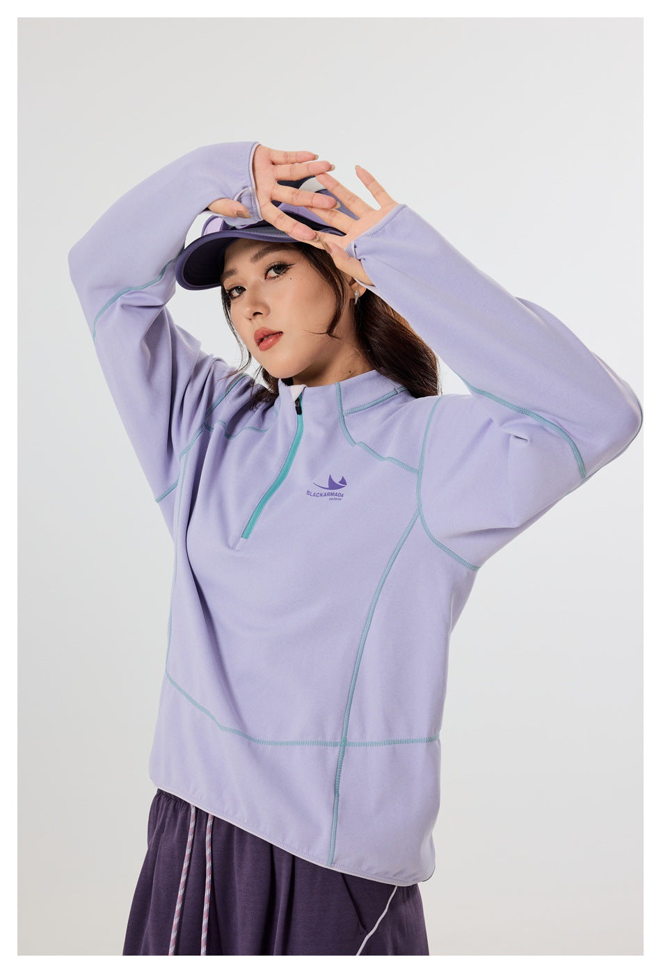 Close view of a lavender quarter-zip pullover showing its teal seams, chest emblem, and long sleeves with thumbholes.