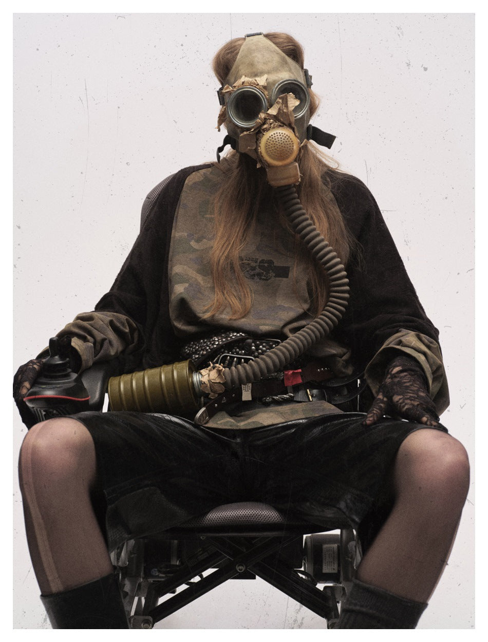 Seated model wearing an olive camouflage raglan top with black sleeves and black shorts, styled with a gas mask.