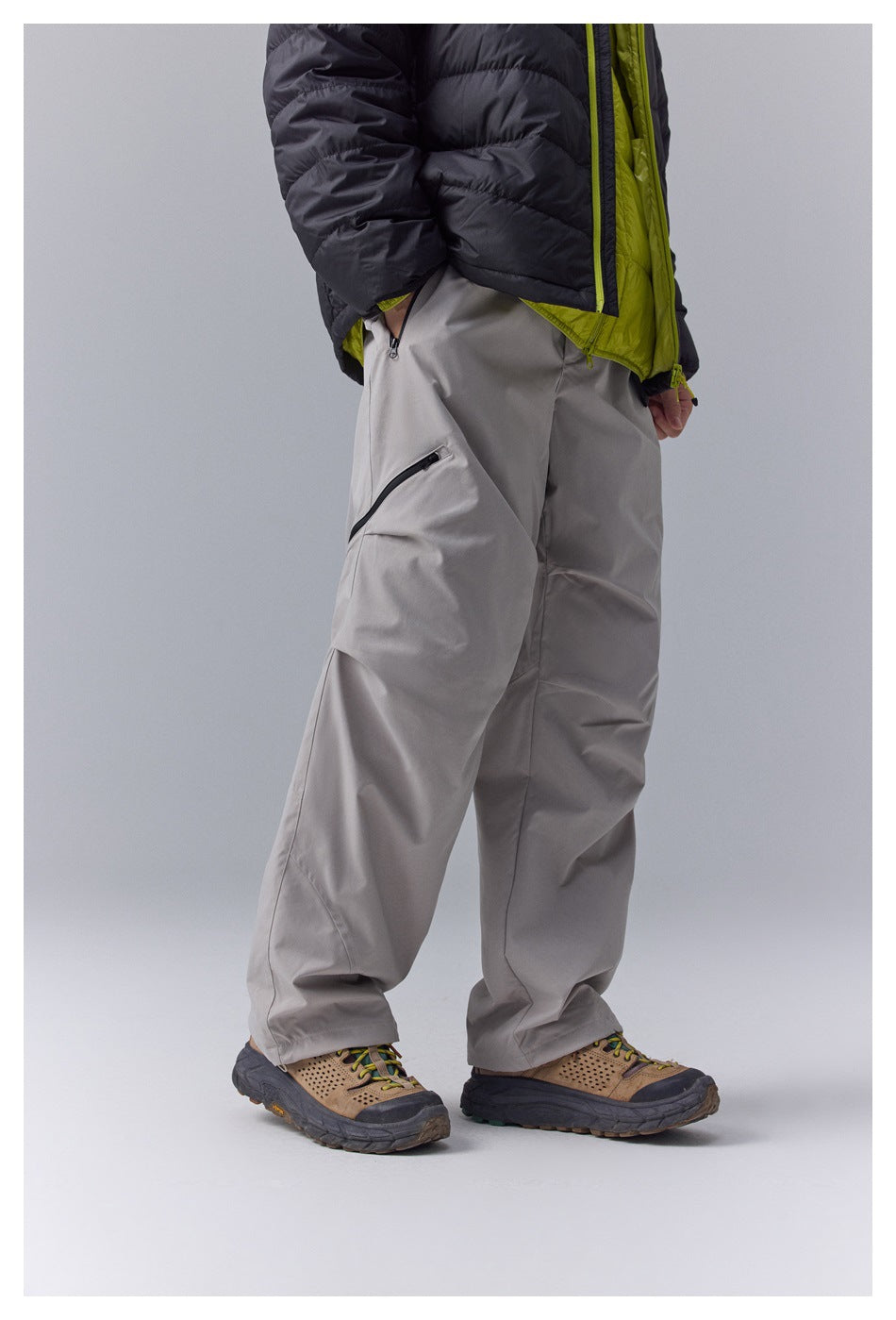 Cropped model wearing loose gray pants with a diagonal thigh zipper, paired with a black and lime jacket and hiking shoes.