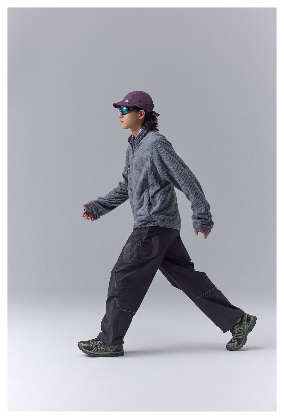 Side view of a model wearing a gray textured zip jacket and loose black cargo pants while walking.