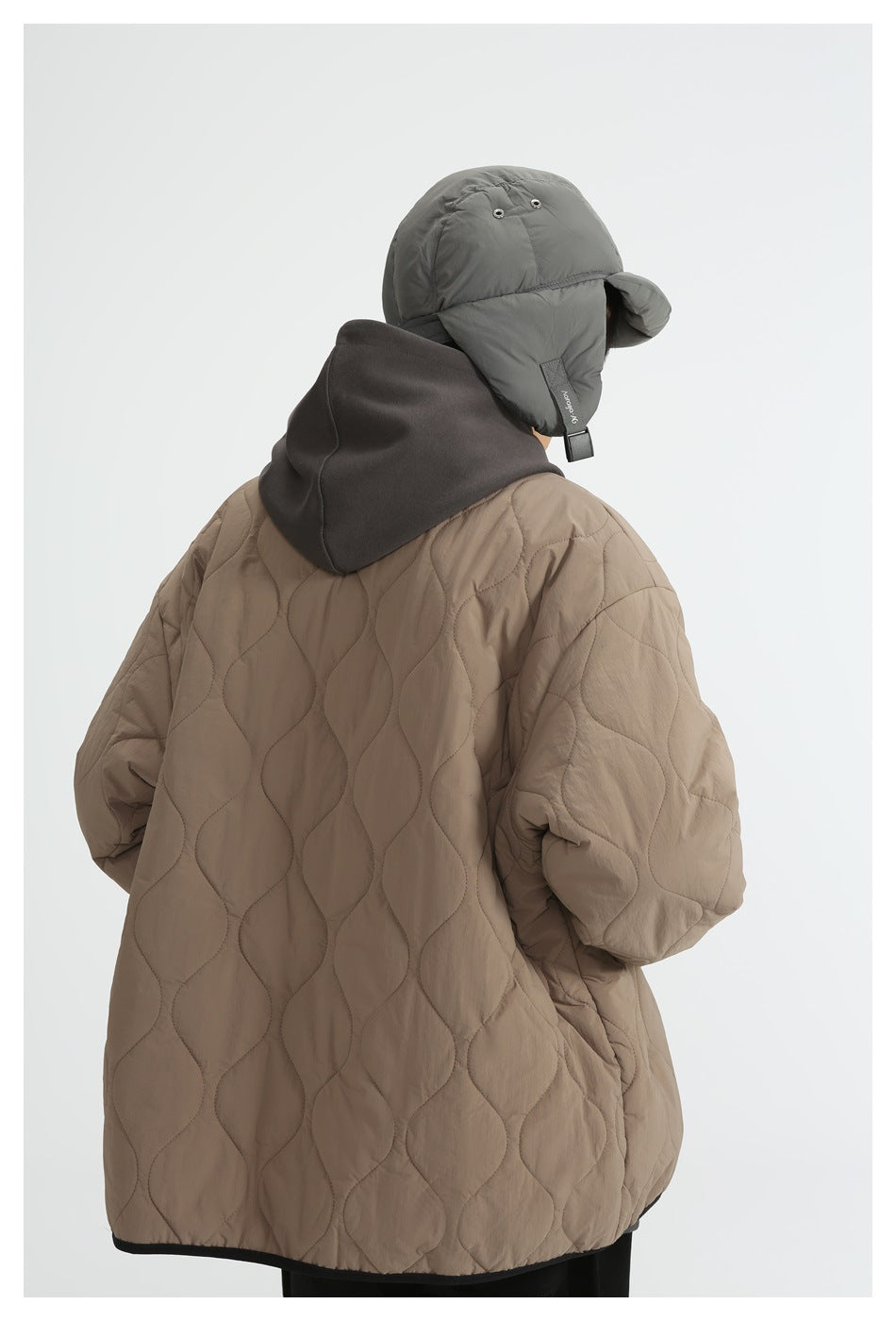 Rear view of a model wearing the taupe quilted jacket, showing its long wavy quilting and black-trimmed hem.
