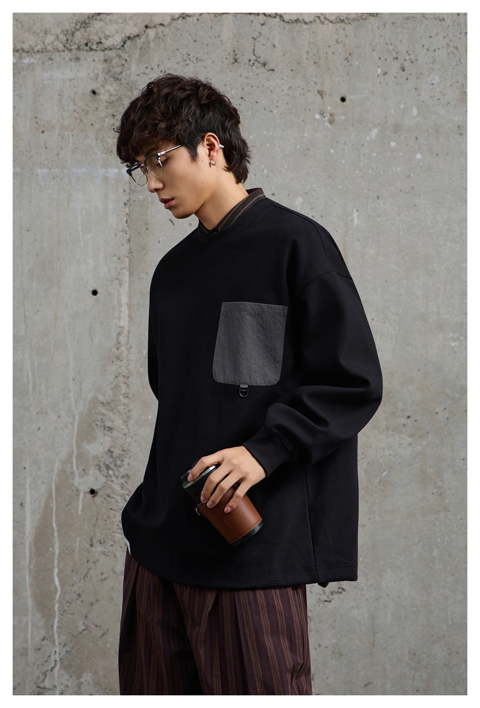 Model in the black crewneck sweatshirt with gray flap pocket, holding a cup, with striped trousers