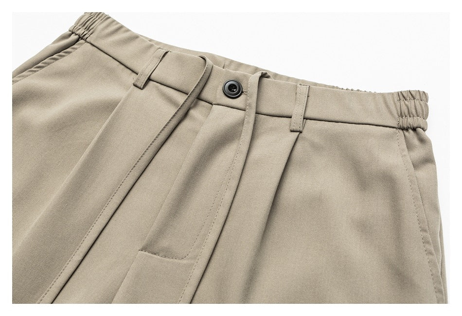 Close-up of taupe trousers showing the front waistband, button, belt loops, and elasticated sides.