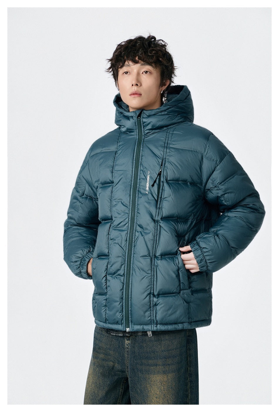 Model wearing a zipped teal hooded quilted puffer jacket with hands in pockets and faded jeans, front view