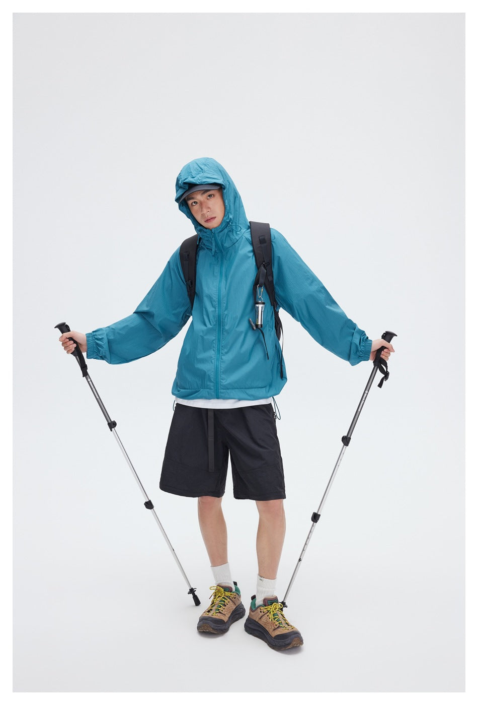 Front view of a person wearing a teal hooded zip jacket and black shorts with a backpack and trekking poles.