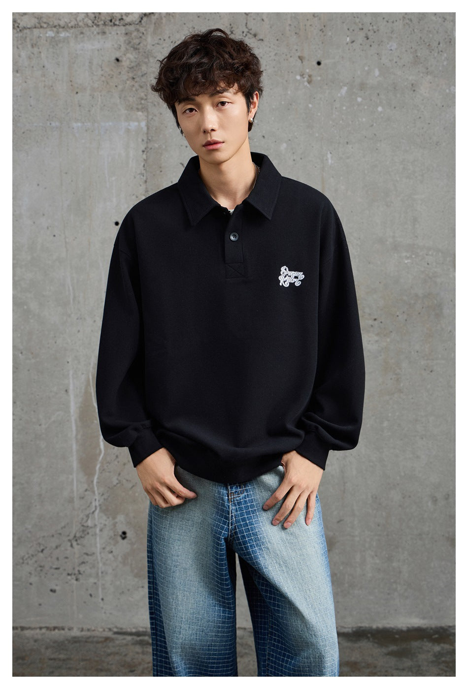 Model wearing a black oversized collared pullover with white chest embroidery and wide blue checked jeans.