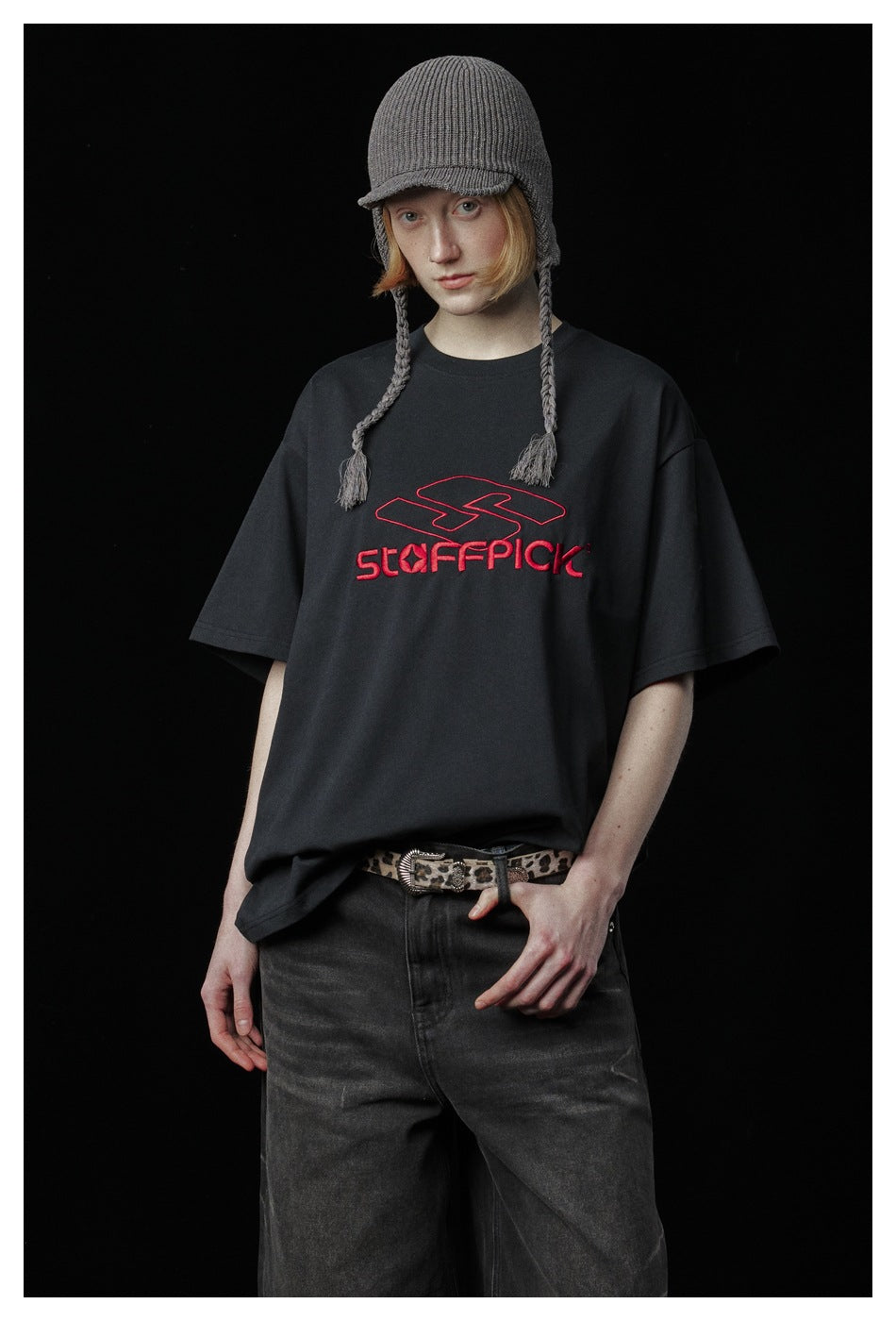 Model wearing a black T-shirt with red chest lettering and a gray knit cap with braided ties.