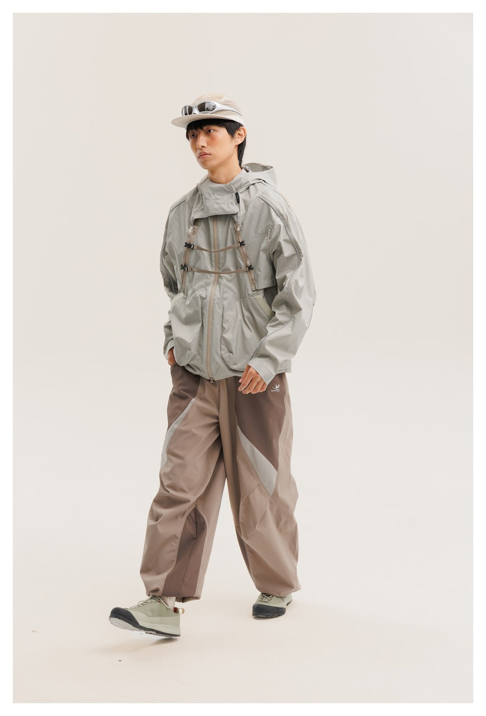 Model in a light gray hooded jacket with chest straps and pockets, two-tone taupe pants and a pale cap.