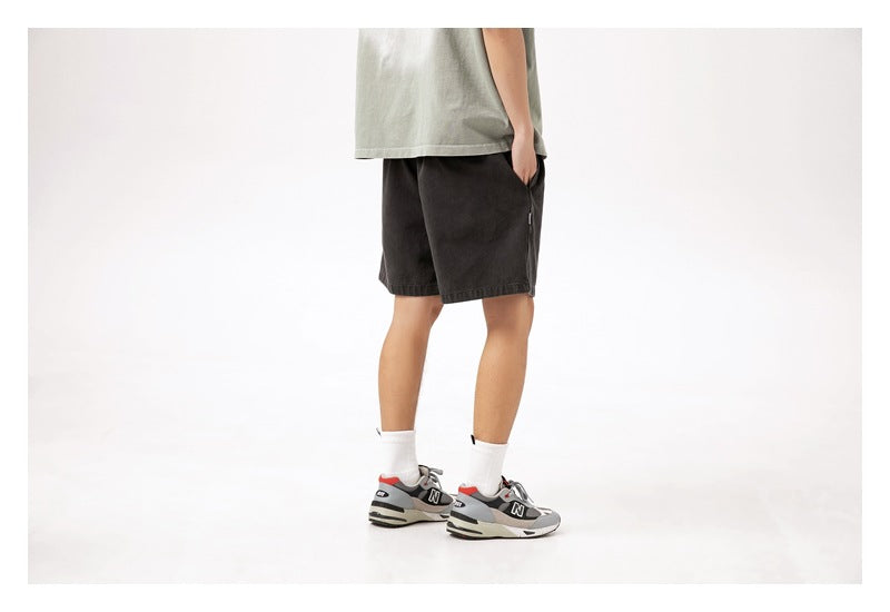Rear three-quarter view of a model wearing charcoal shorts, a light gray-green shirt, white socks and sneakers.