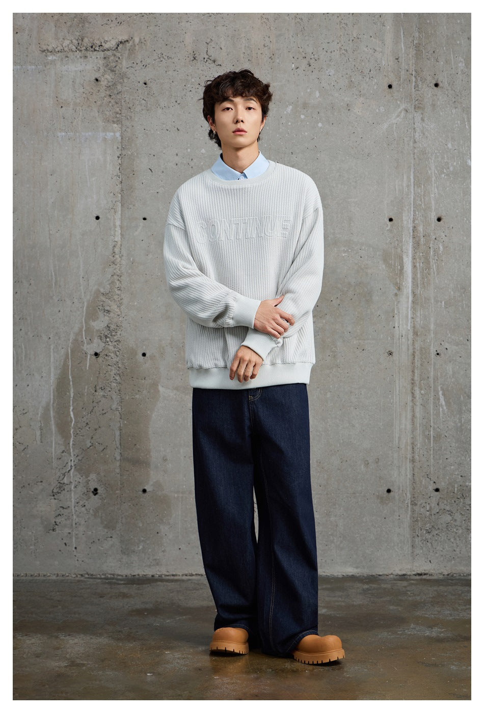 Full-length view of a model wearing an off-white textured sweater, pale blue shirt, and dark jeans.