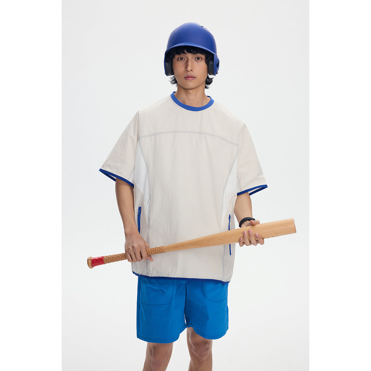 Model wearing an oversized ivory shirt with blue trim and bright blue shorts, holding a baseball bat.