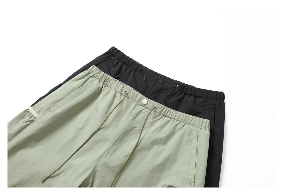 Two pairs of light sage and black utility pants overlap, showing elastic waistbands, drawcords, and front pockets.