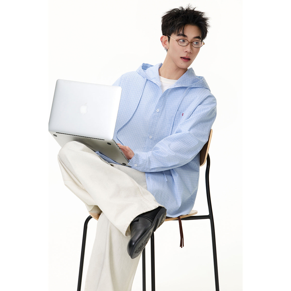Seated model wearing an oversized light blue checked hooded shirt, cream trousers and black shoes, using a laptop.