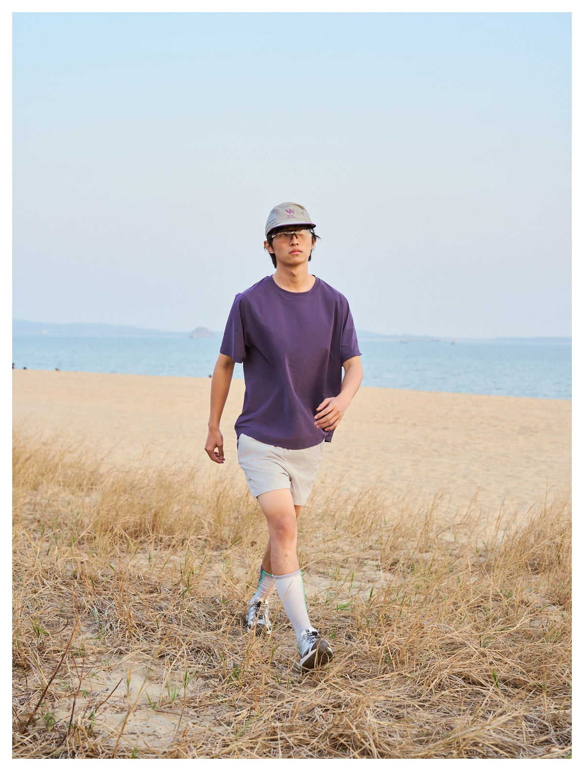 Man wearing a plain purple short-sleeve T-shirt and light shorts, walking through beach grass.