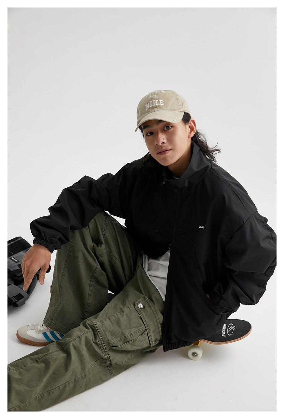 Seated model wearing a black zip-front jacket with a stand collar and elastic cuffs, styled with olive cargo pants.