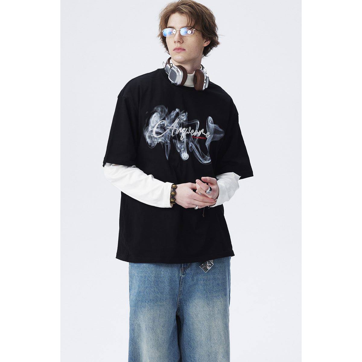 Model wearing an oversized black graphic T-shirt over a white long-sleeve shirt, with loose blue jeans.