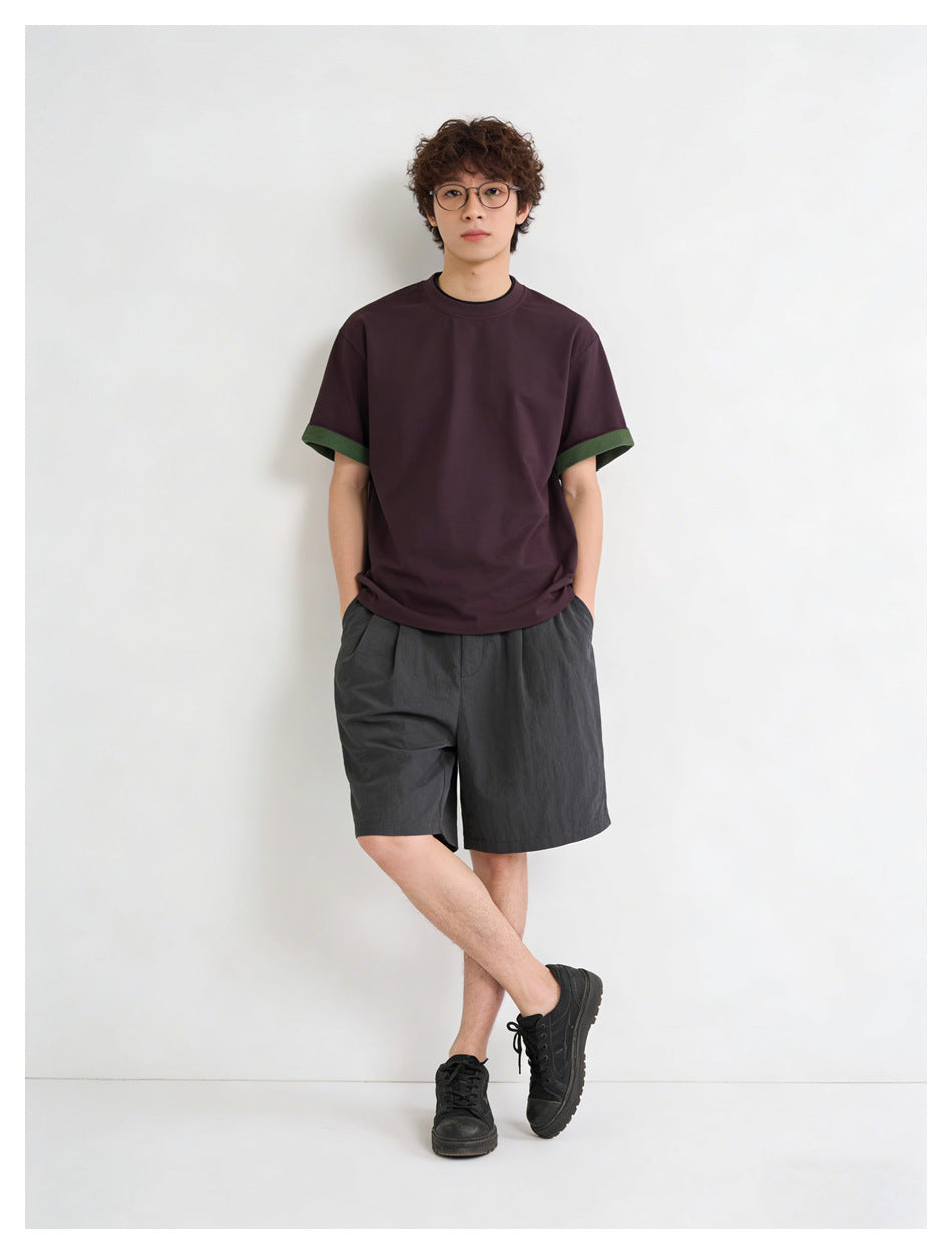 Model wearing a dark plum crew-neck T-shirt with green sleeve cuffs and charcoal shorts, shown full length.