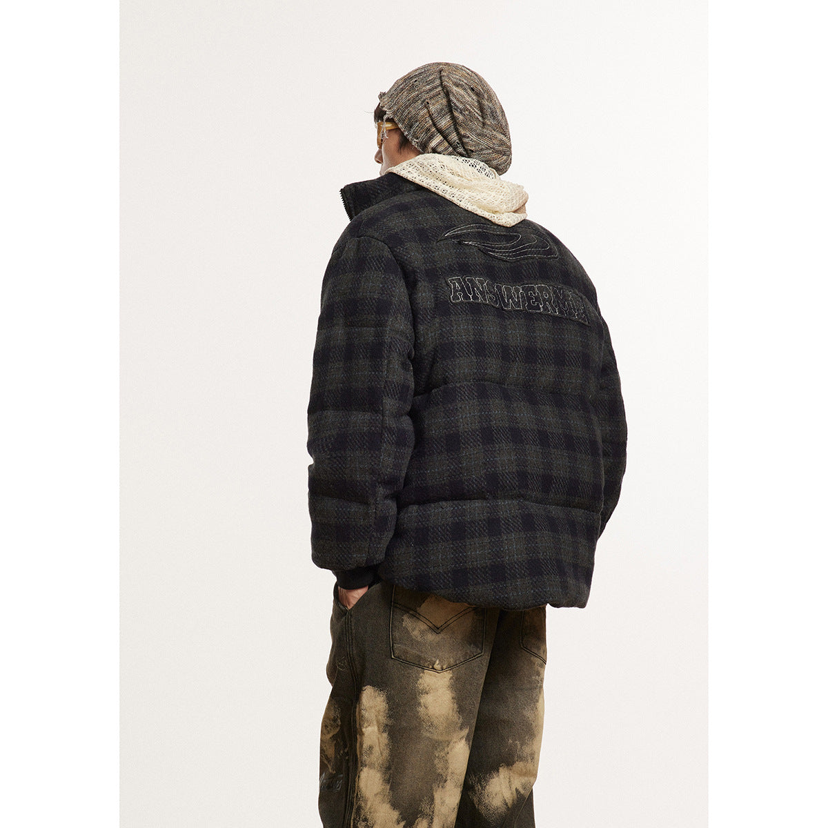 Rear three-quarter view of a dark plaid puffer jacket with outlined lettering on the back, worn with mottled pants.