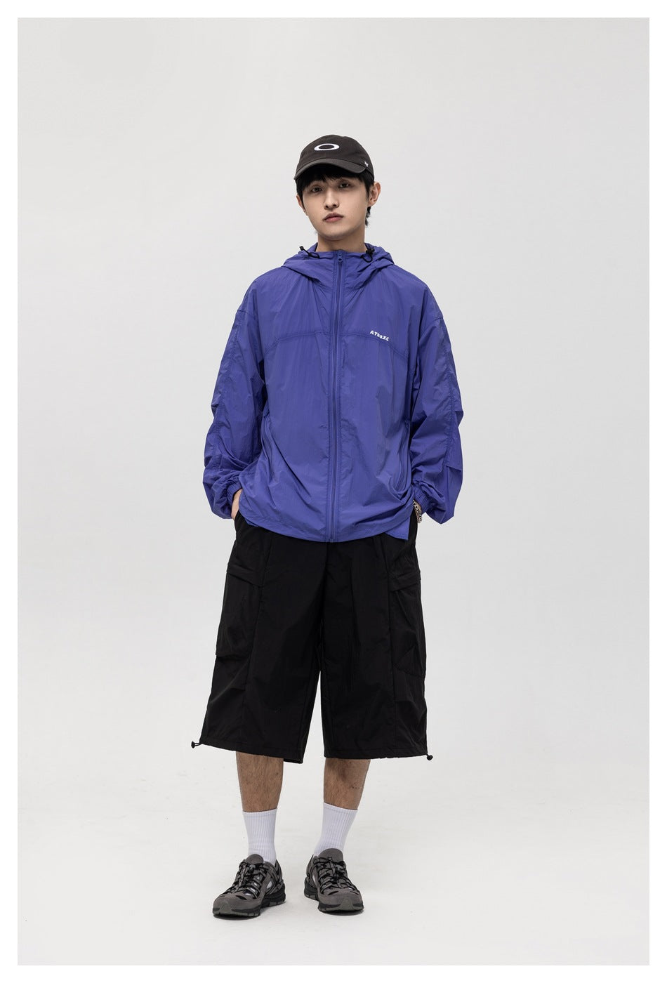 Model wearing a blue hooded zip jacket with black cargo shorts, cap and gray sneakers, front view