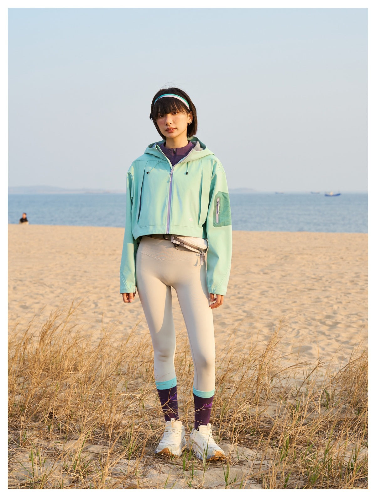 Full-length beachside view of a model wearing a mint hooded cropped jacket, cream leggings, and purple socks.